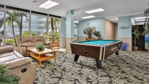 Une table de billard dans une pièce dotée de grandes fenêtres, d’un canapé et d’une petite table sur laquelle est posée une plante.