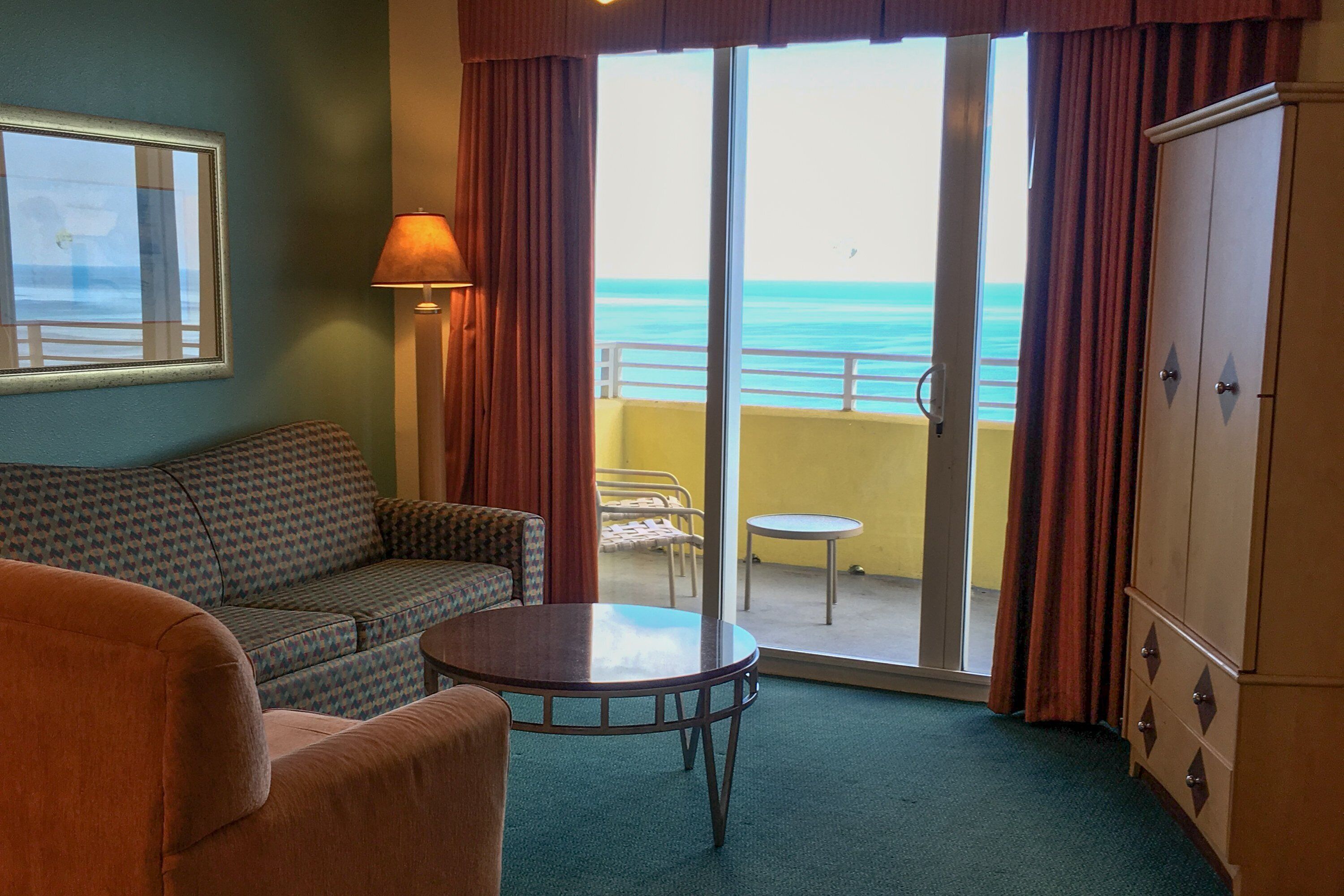 A room with a sofa, a table, a lamp, a wardrobe, and a view of the ocean through a sliding door.