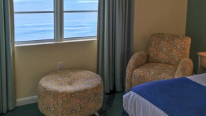 A hotel room with a view of the ocean, a patterned armchair, a round ottoman, and a bed with a blue blanket. - Ocean Walk 2424a (Daytona Beach)