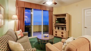 A hotel room with a sofa, armchair, and a small table with a bowl. There is a television on a wooden cabinet and a ceiling fan. - Ocean Walk 2424a (Daytona Beach)