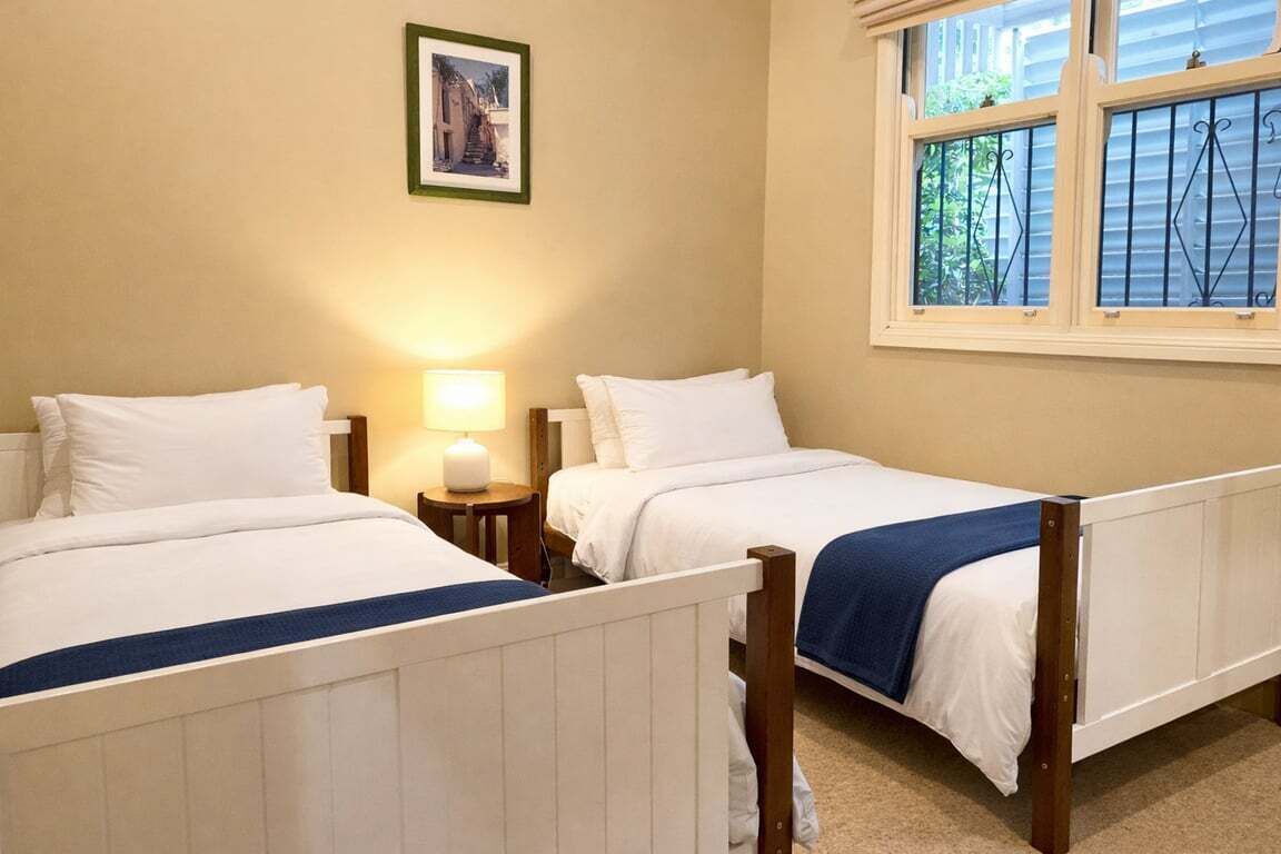 Camera da letto con due letti, un comodino con lampada, un quadro appeso al muro e una finestra vista sul verde.