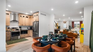 A modern kitchen with stainless steel appliances, wooden cabinets, and a dining area with orange chairs and a black table set for six. - Modern Group Oasis: Hot Tub, Arcade & Fire Pit (St. Louis)