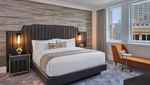 Una camera d'albergo moderna con un grande letto, due comodini con lampade, un divano e vista sulla città attraverso un'ampia finestra.
