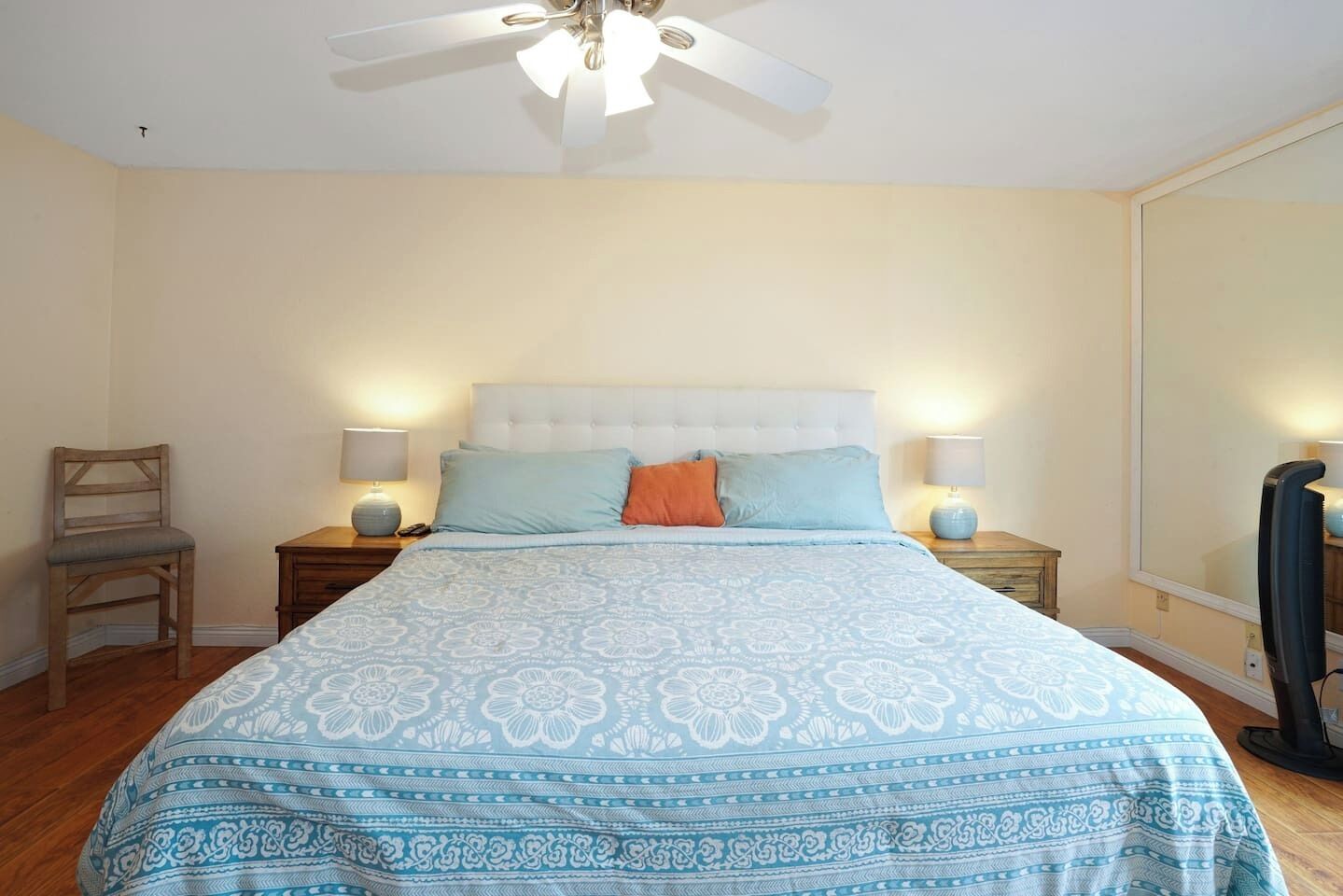 A bedroom with a bed, two bedside tables, a chair, a ceiling fan, and a large mirror.
