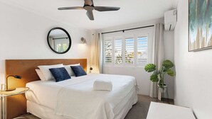 Une chambre d’hôtel avec un lit, un ventilateur de plafond, un miroir rond, une lampe de chevet, une plante en pot et une fenêtre avec des stores.