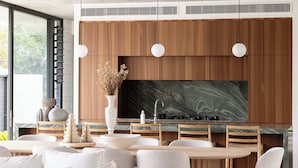 Uma cozinha moderna com armários de madeira, uma televisão grande e uma sala de jantar com cadeiras claras e uma mesa branca.