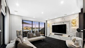 A modern living room with a large flat-screen TV, comfortable seating, and a view of the cityscape through large windows. - Elegant Waterfront Escape – Point Cook

Relax with the whole family. (Point Cook)
