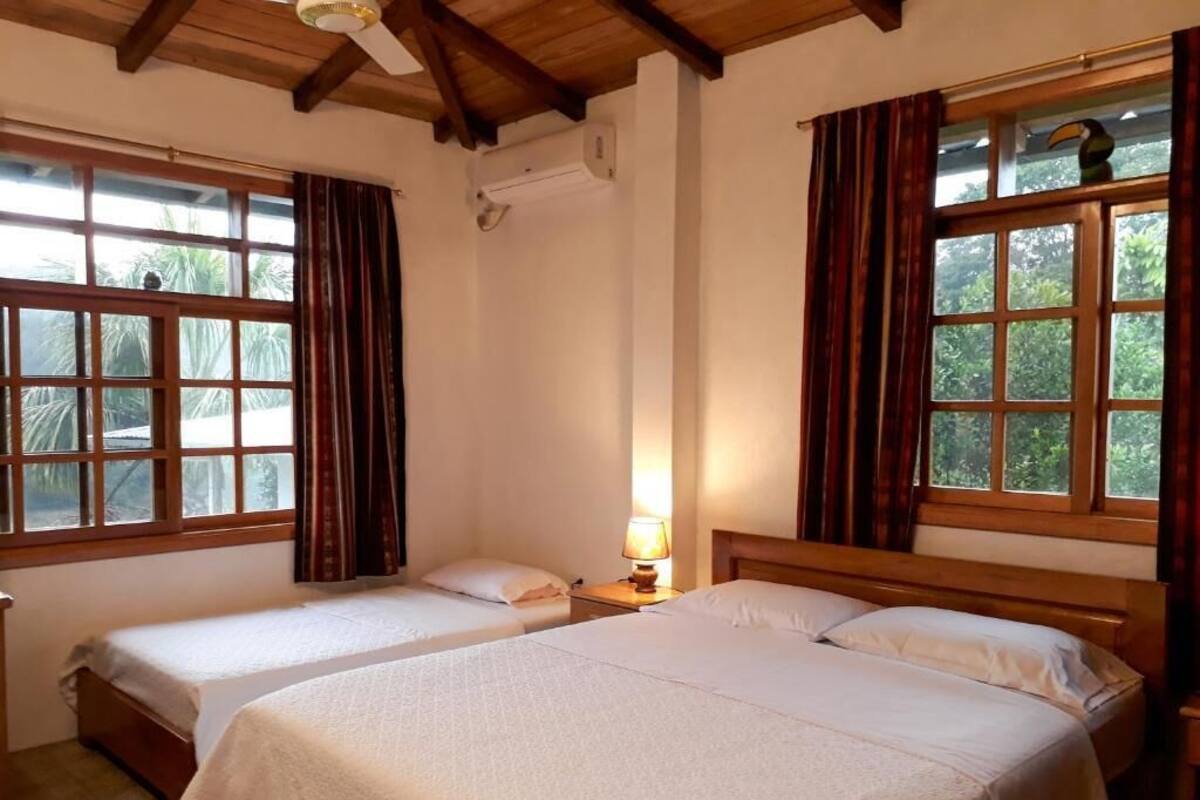A room with two beds, a window with curtains, a ceiling fan, and a nightstand with a lamp.