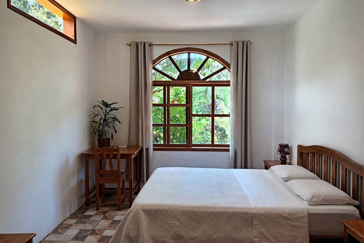 A bedroom with a bed, a desk, a chair, a potted plant, and a window with a view of greenery.
