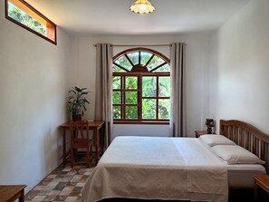 A bedroom with a bed, a desk, a chair, a potted plant, and a window with a view of greenery.