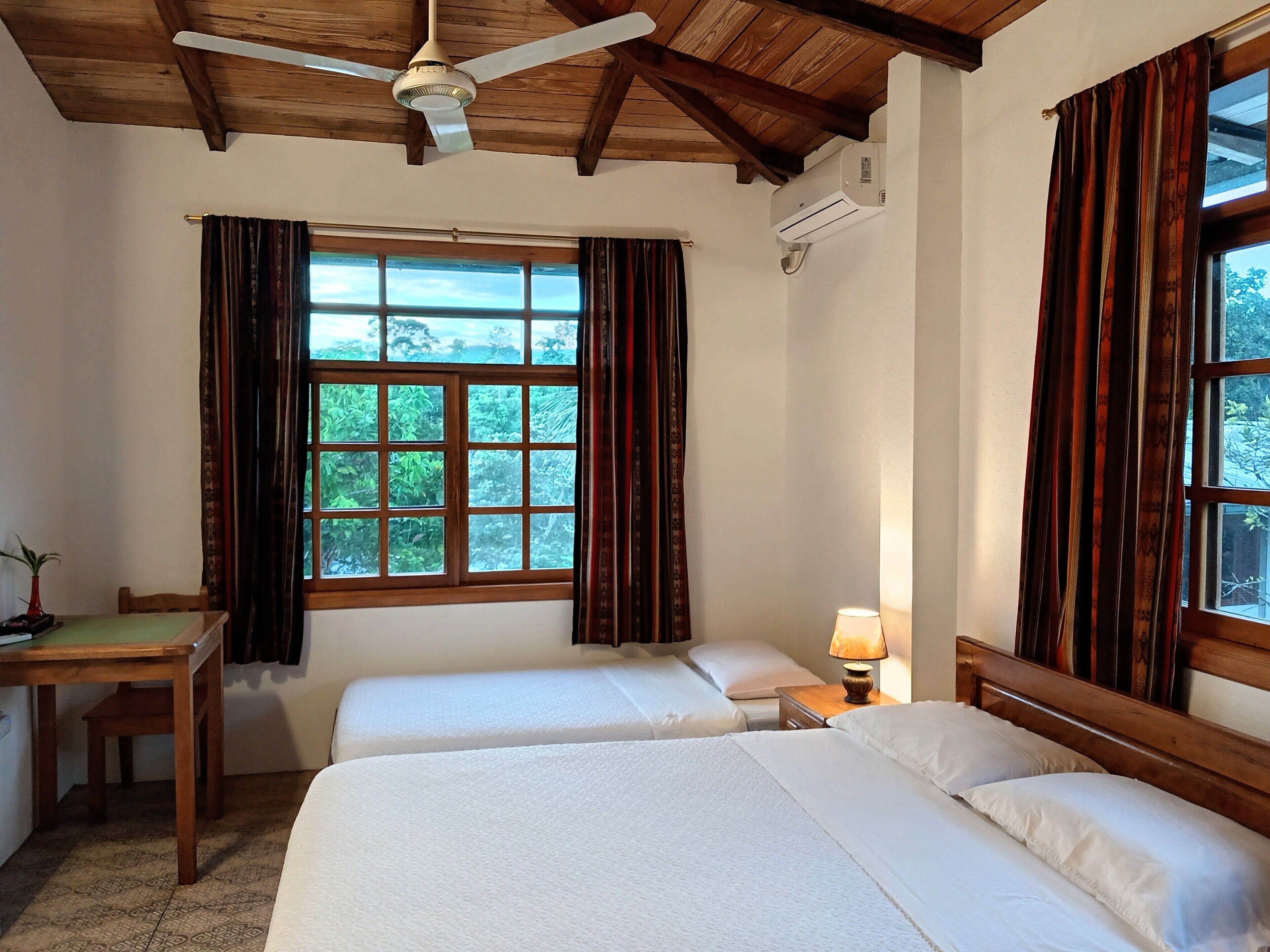 A room with two beds, a desk, a ceiling fan, and a window with curtains.