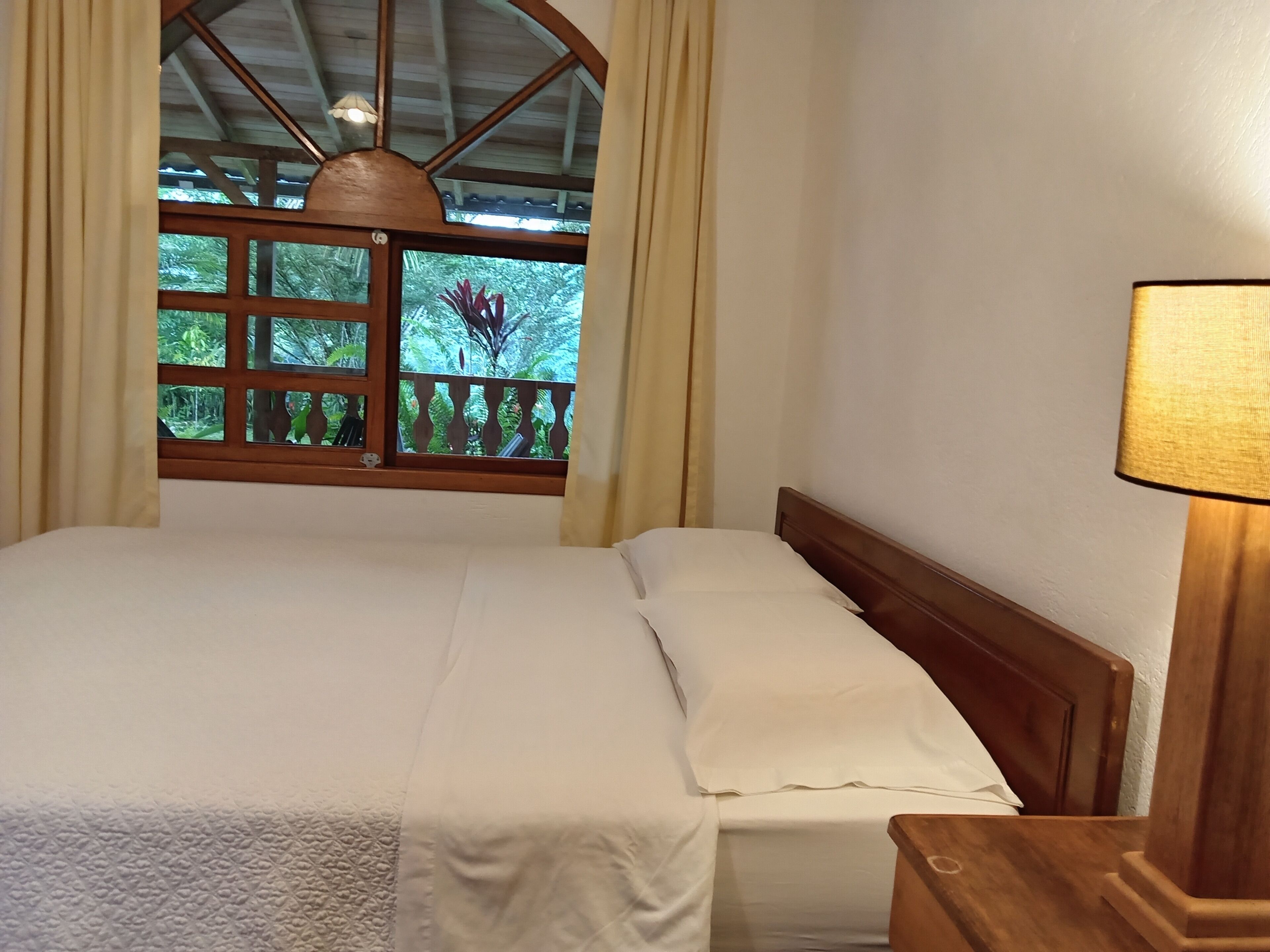 A neatly made bed with a wooden headboard, a window with a view of greenery, and a lamp on the bedside table.