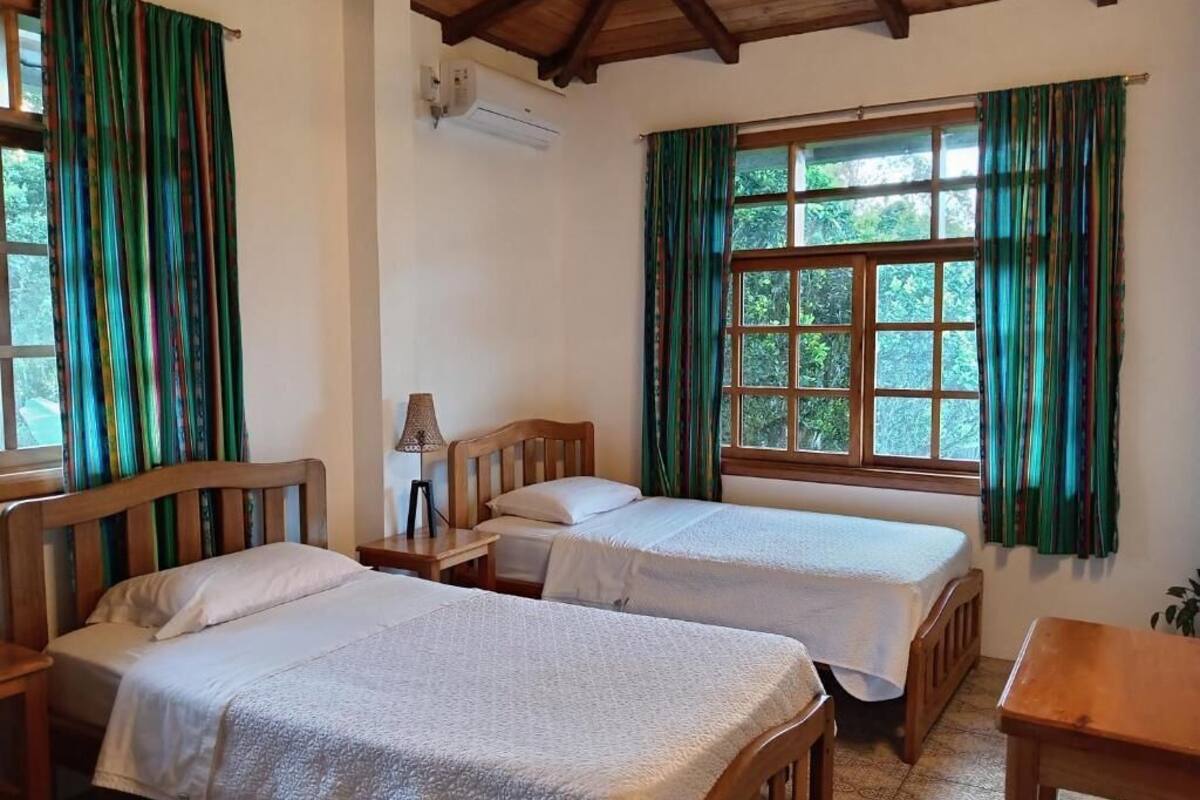 A room with two wooden beds, a wooden table, a ceiling fan, and a window with curtains.