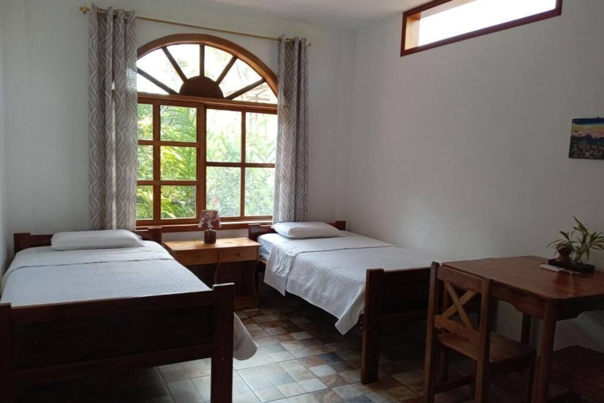 A room with two wooden beds, a small wooden table, a window with curtains, and a tiled floor.