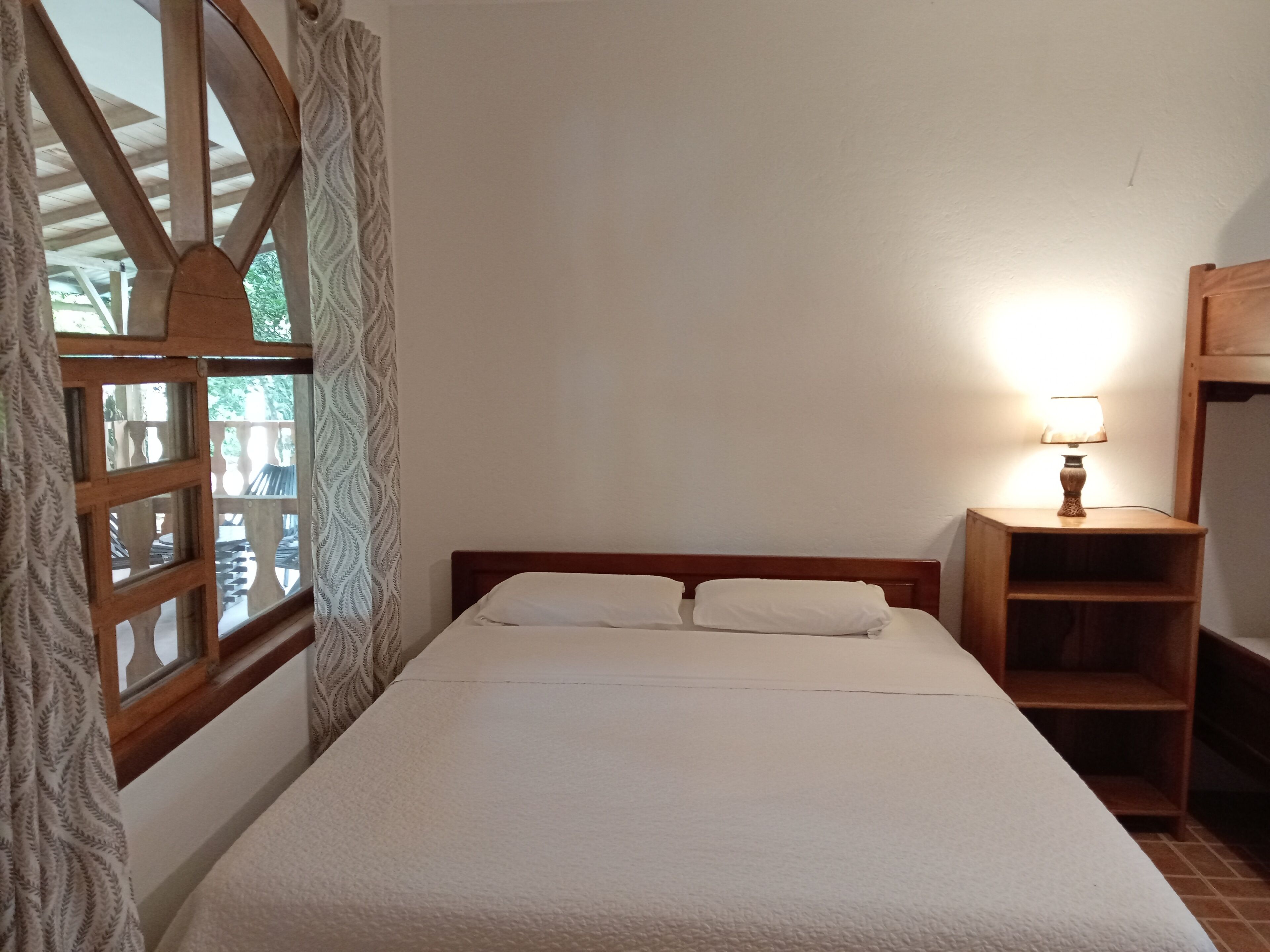 Un dormitorio con cama de madera, ropa de cama blanca, una mesita de noche con lámpara y una ventana con cortinas estampadas.