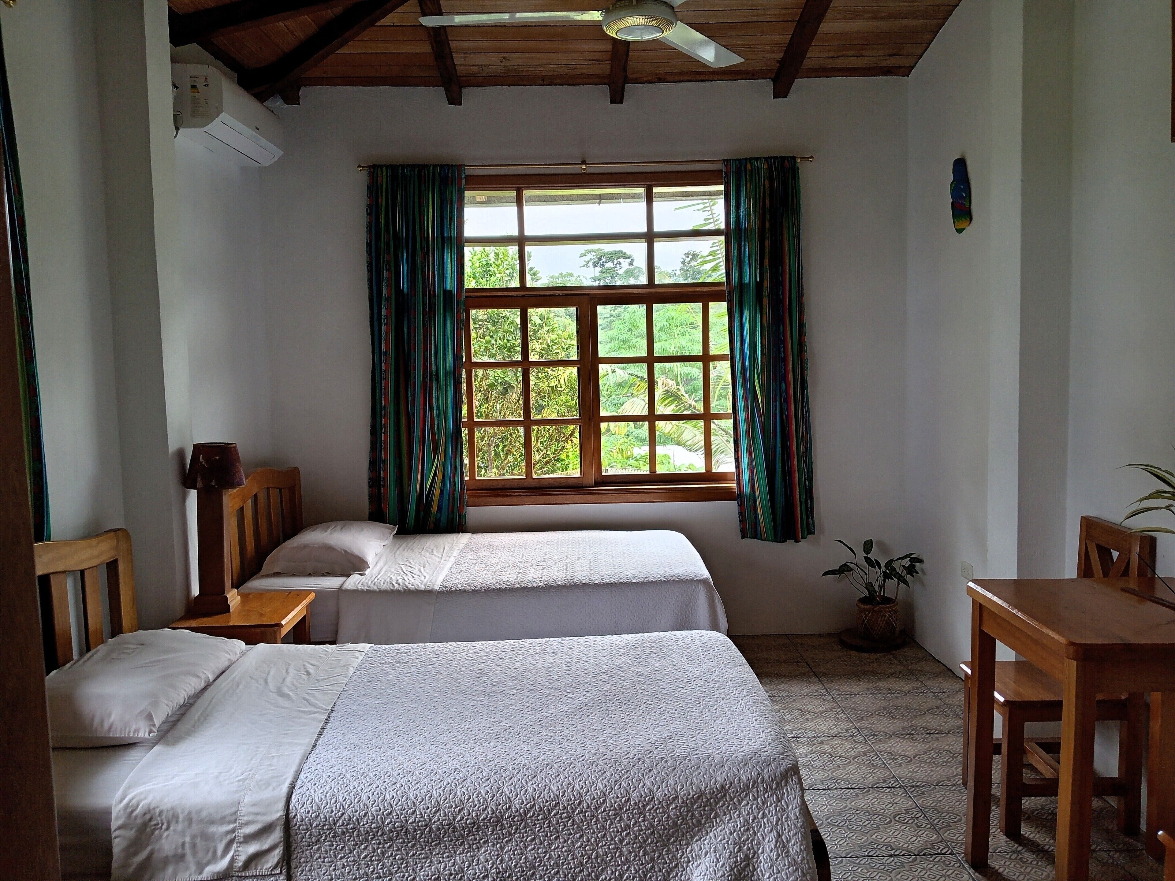 A room with two beds, a wooden ceiling, a window with curtains, a ceiling fan, and a small table.