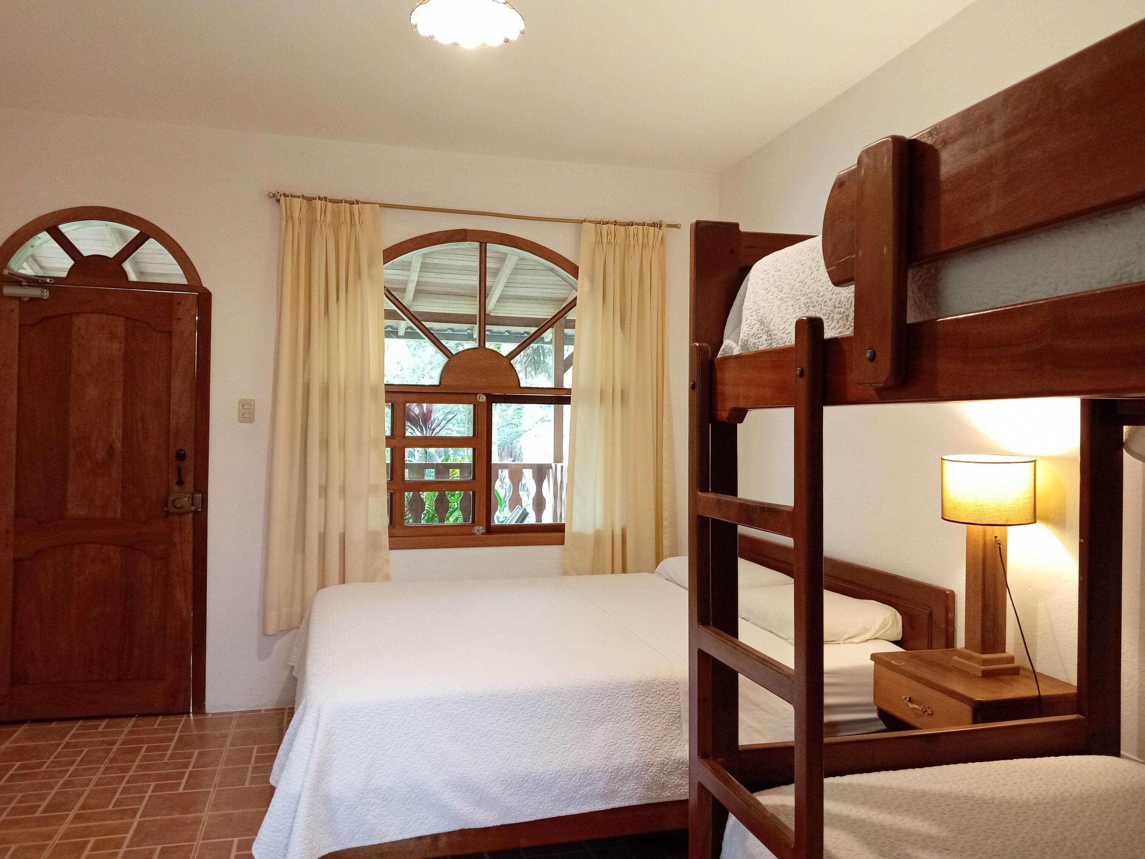 Una habitación con una cama litera, una cama individual, una puerta de madera, una ventana con cortinas y una lámpara de noche.