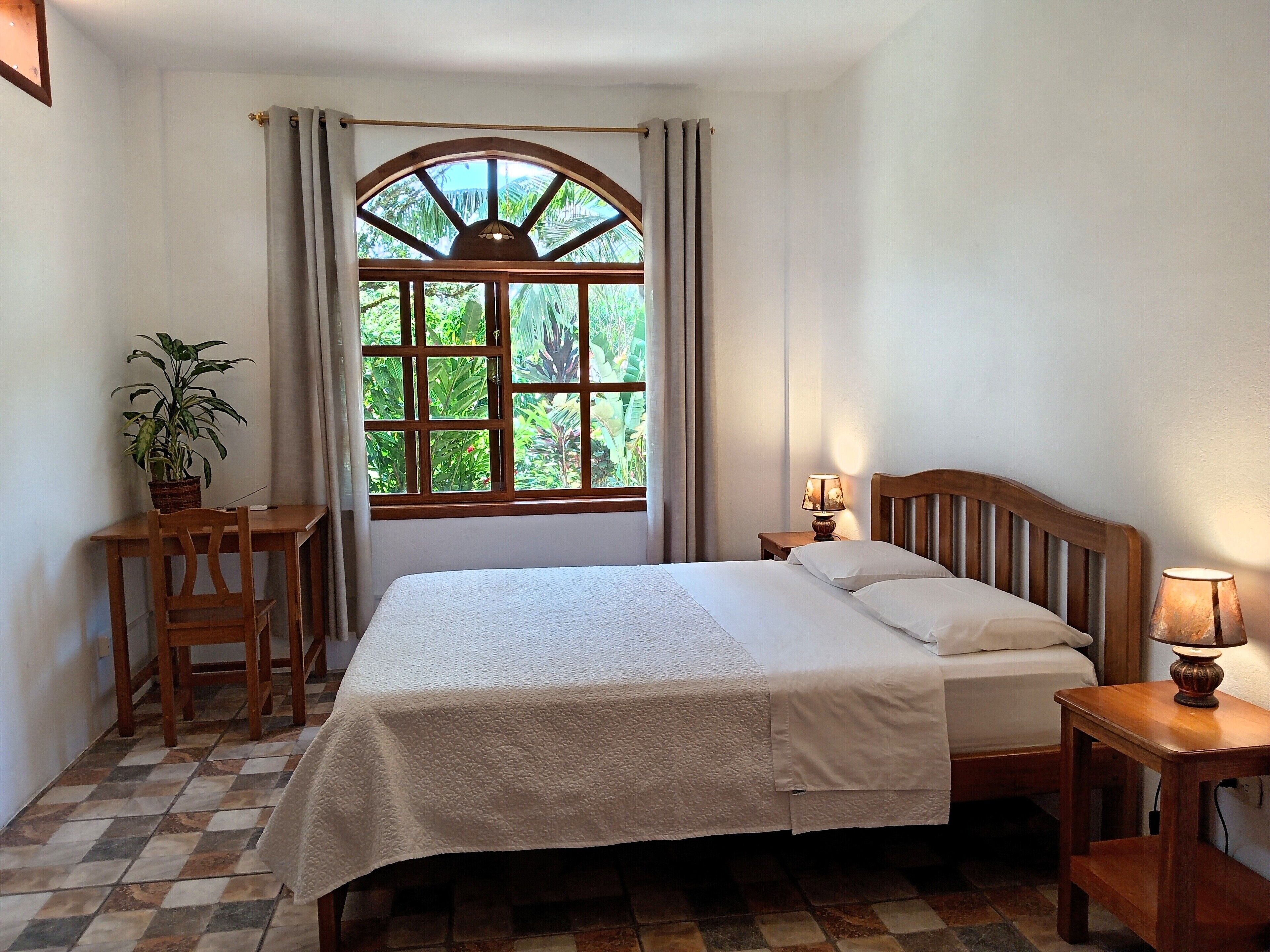 A bedroom with a bed, a nightstand, a lamp, a chair, a potted plant, and a window with curtains.