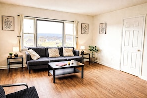 A living room with a sofa, coffee table, and two side tables. There are framed pictures on the walls and a door in the background. - Spacious 5BR Lake View Near Cornell Sleeps 9 • Accessible • Perfect for Groups (Ithaca)