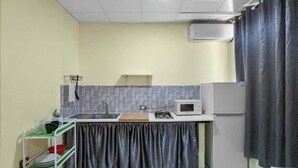 Une cuisine compacte équipée d’un four à micro-ondes, d’un évier et d’un petit réfrigérateur. On y trouve une étagère verte où sont rangés de la vaisselle, et un rideau noir qui recouvre la fenêtre.