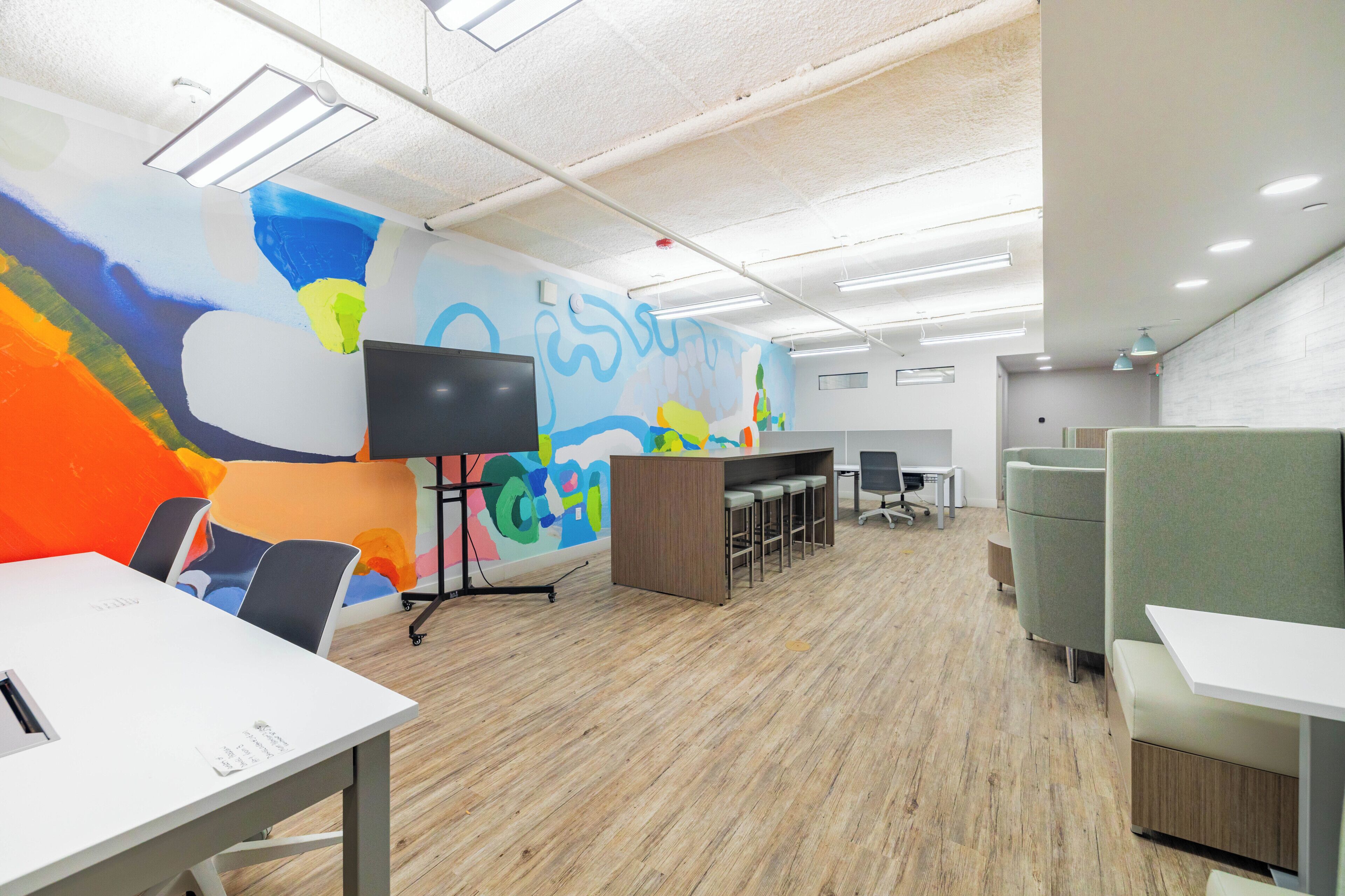 A modern office space with a large colorful mural, a TV on a stand, and a long table with chairs.