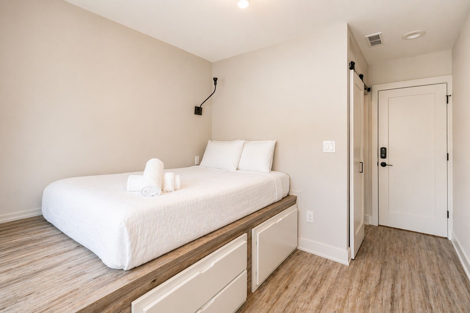 Une chambre à coucher avec un lit, une table de chevet, une lampe fixée au mur, une porte et un plancher en bois.