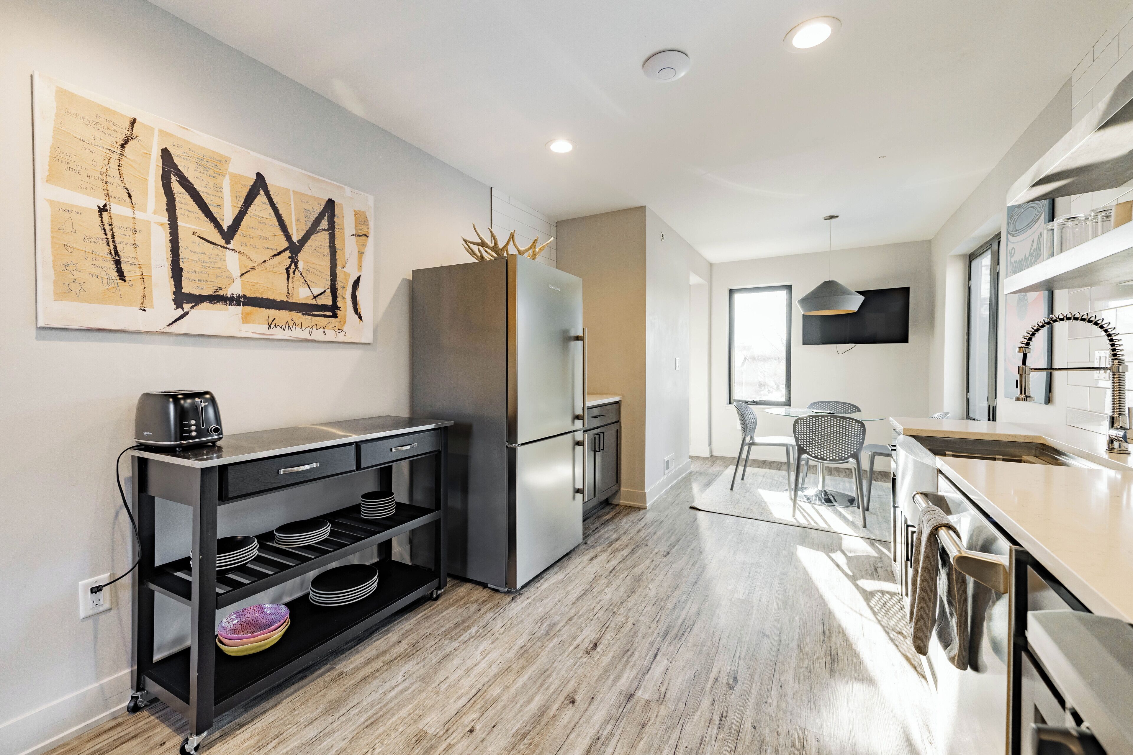 A modern kitchen with a stainless steel refrigerator, a black cart with dishes, and a large abstract painting on the wall.