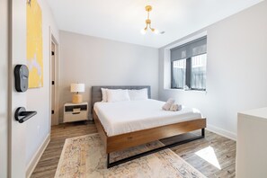 A modern bedroom with a wooden bed, a nightstand, a lamp, a wall-mounted light fixture, and a large window. - Stadium Suites (Ann Arbor)
