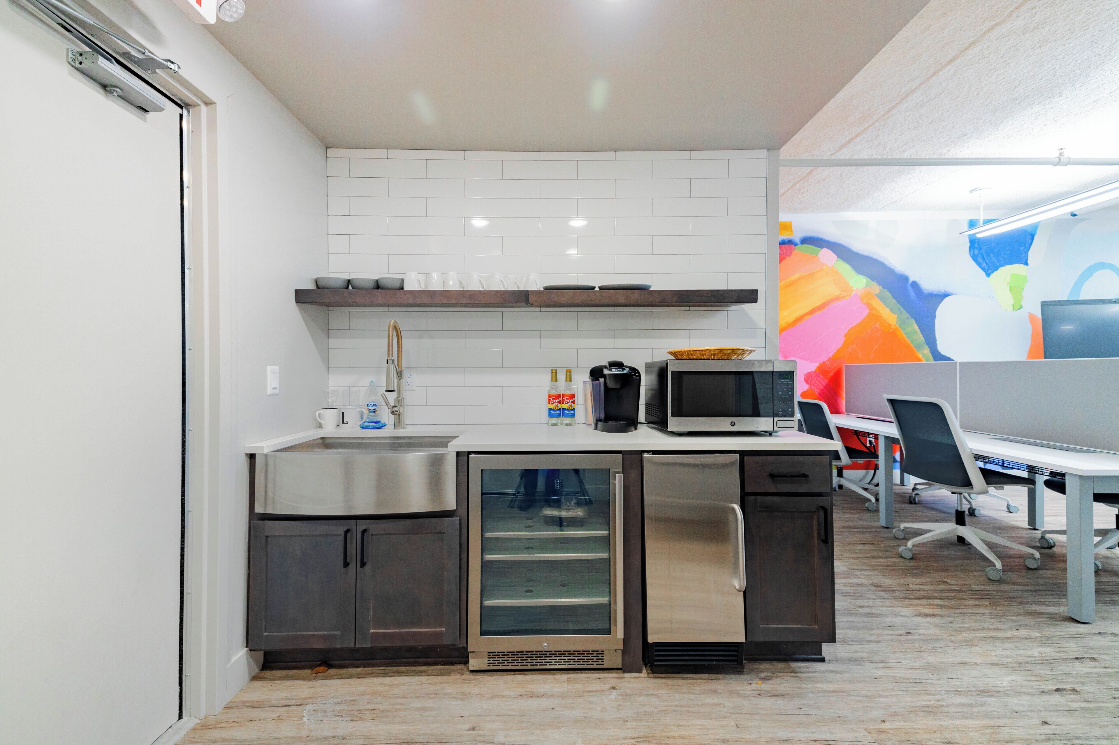 A modern kitchen with stainless steel appliances, a microwave, and a refrigerator. There is a white tiled backsplash and a colorful mural on the wall.