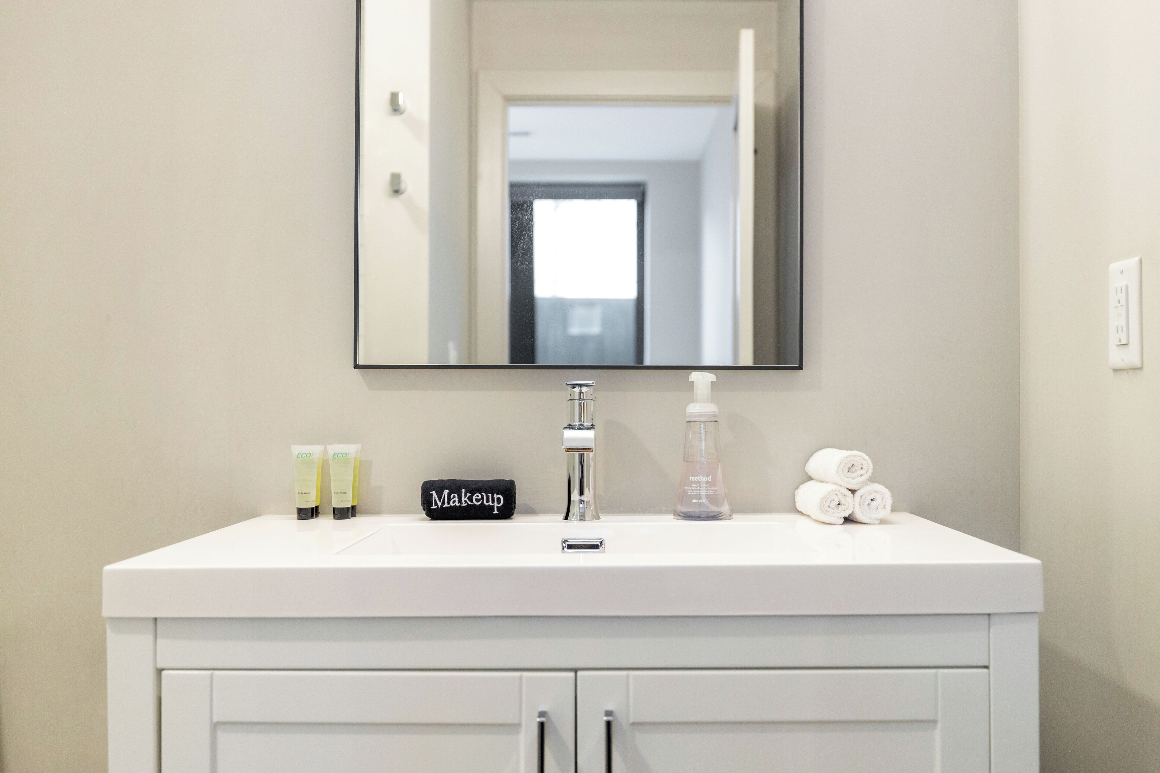 Un tocador de baño blanco con un espejo grande, un lavabo y un armario debajo. Sobre el tocador, hay dos botellas de loción, un organizador de maquillaje y una botella de agua.