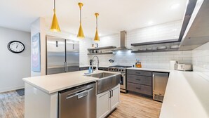 A modern kitchen with stainless steel appliances, a dishwasher, and a sink. There are yellow pendant lights, a clock on the wall, and a tiled backsplash. - Stadium Suites (Ann Arbor)
