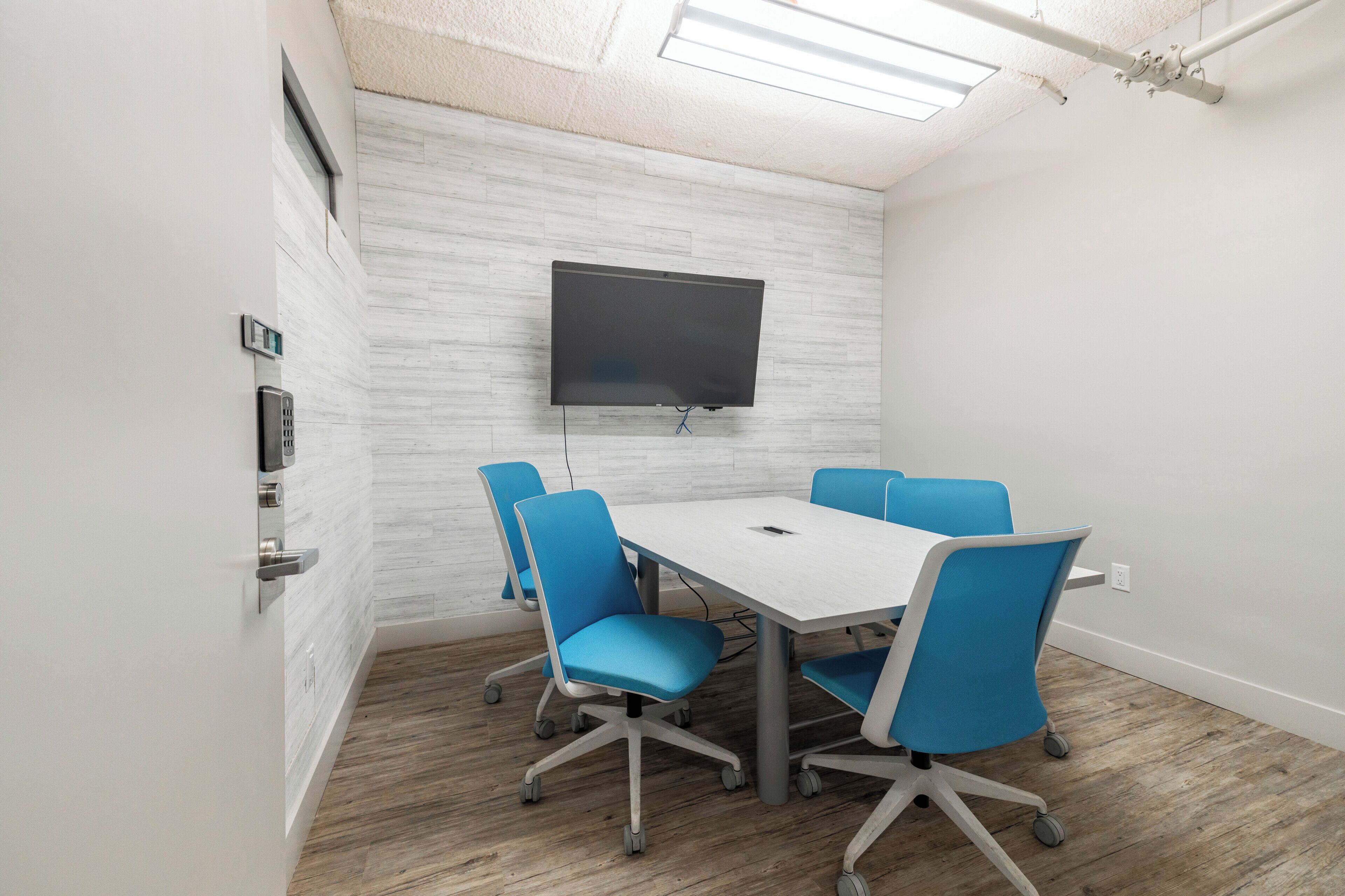 A conference room with a white table, blue chairs, a mounted TV, and a wooden floor.