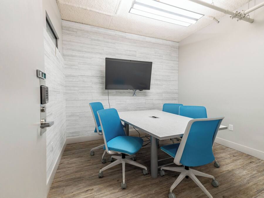 A conference room with a white table, blue chairs, a mounted TV, and a wooden floor.