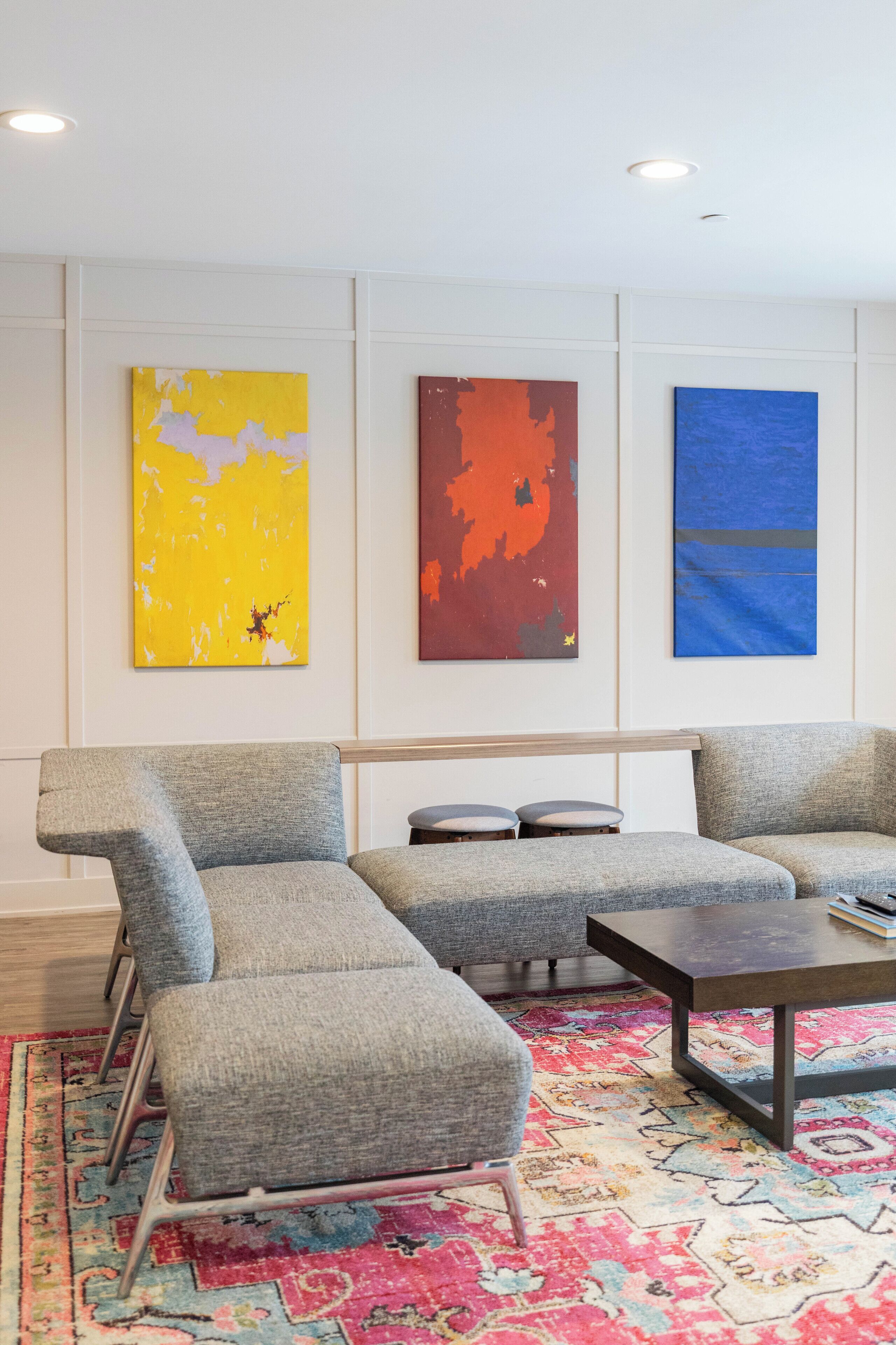 A modern living room with a gray sectional sofa, a small coffee table, and a colorful rug. There are three abstract paintings on the wall.