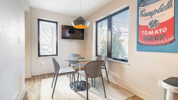 A dining area with a glass table, metal chairs, a hanging pendant light, and a large poster of Campbell's Tomato Soup.