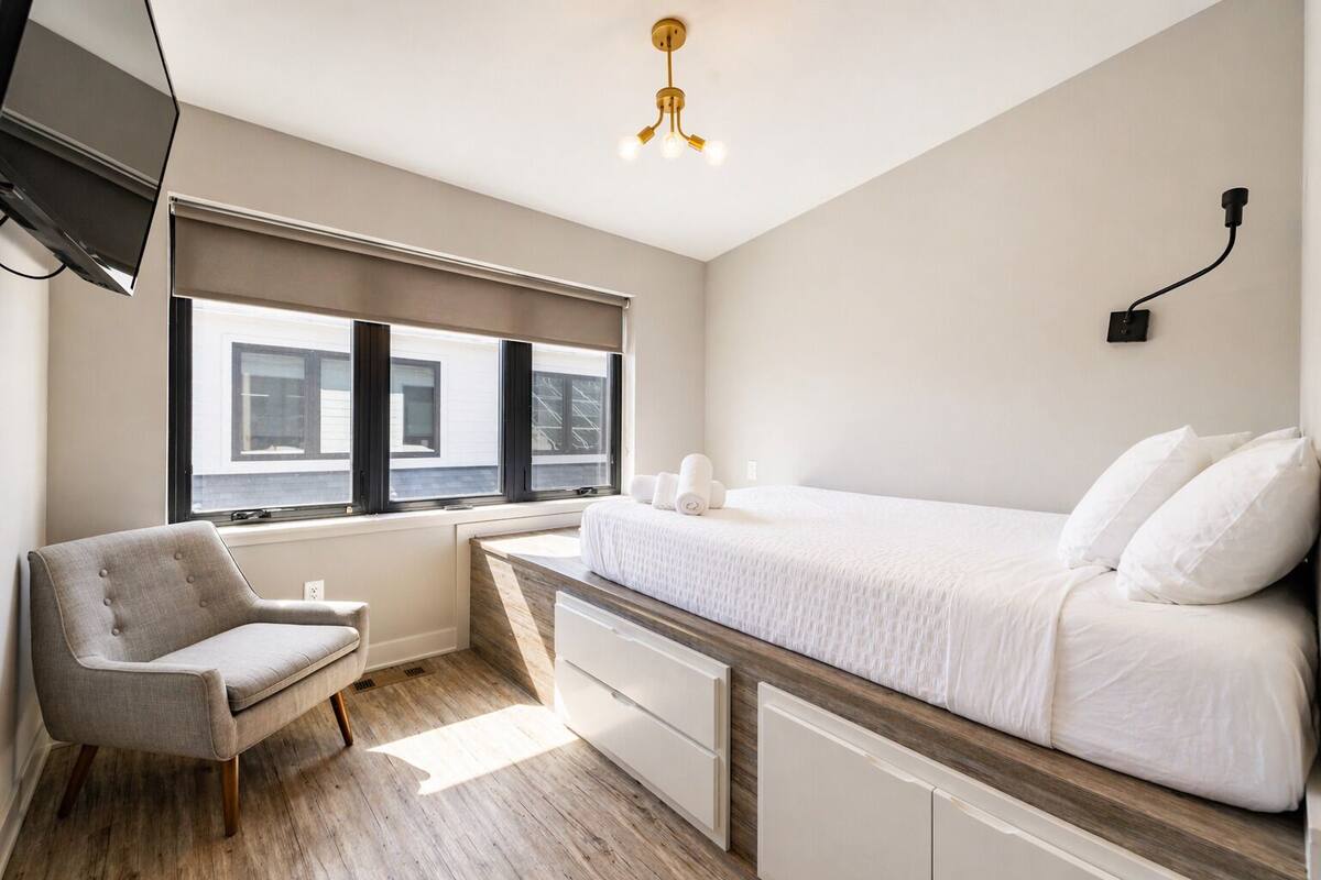 A bedroom with a bed, a chair, a desk, and a window with blinds.