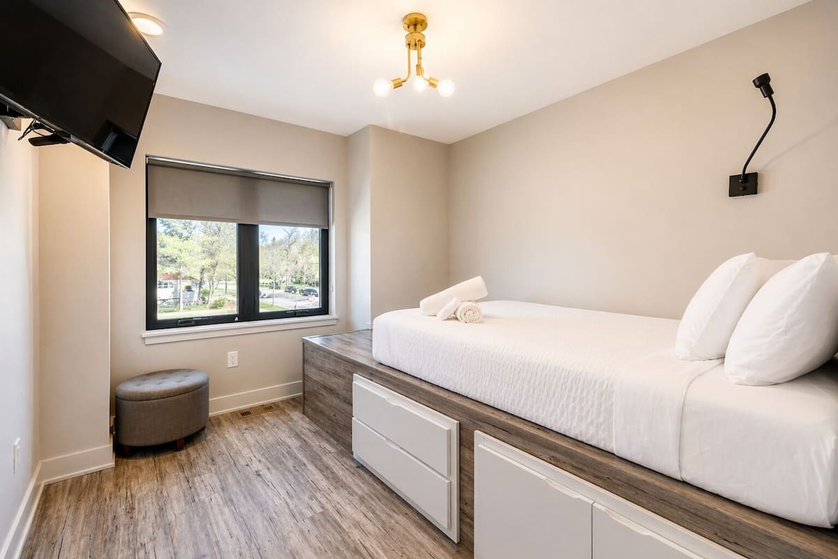 A bedroom with a bed, a window, a television, a bedside table, and a ceiling light fixture.
