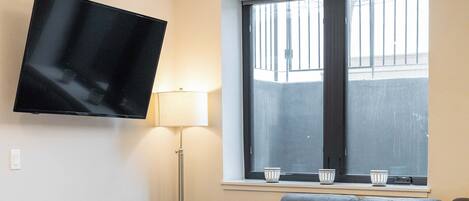 Ein modernes Wohnzimmer mit einem an der Wand befestigten Flachbildfernseher, einem grauen Ecksofa, einem runden Hocker, einem gelben Sessel und einem großen Fenster mit Jalousien.