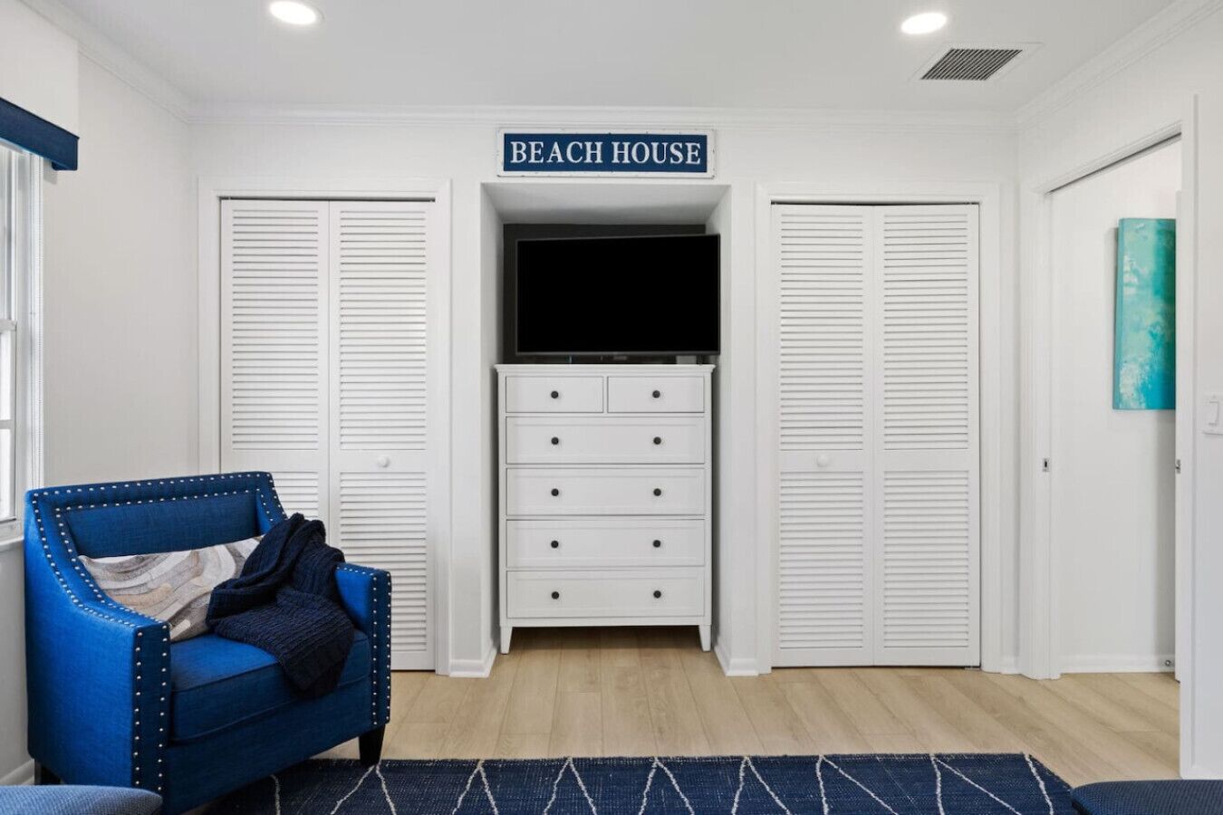 Una habitación con una silla tapizada de azul, un aparador blanco con cajones y un cartel sobre una puerta que dice "BEACH HOUSE".
