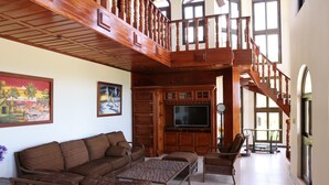 A spacious living room with a large wooden staircase, a flat-screen TV, wicker furniture, and large windows. - Beautiful home and gardens near the beach (Ojochal)