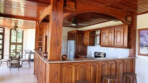 A kitchen with wooden cabinetry, a central island with stools, and a dining area with chairs and a table. - Beautiful home and gardens near the beach (Ojochal)
