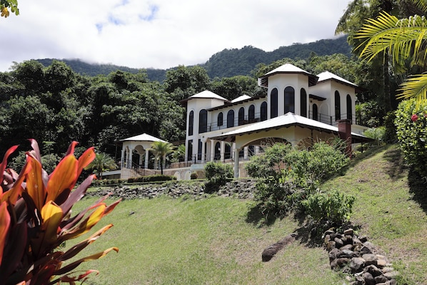 A white building with arched windows and a balcony, surrounded by lush greenery and a hillside.