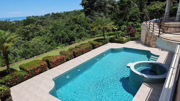 A rectangular swimming pool with a hot tub, surrounded by a landscaped garden with red flowers and palm trees, and a view of a forested area. - Beautiful home and gardens near the beach (Ojochal)