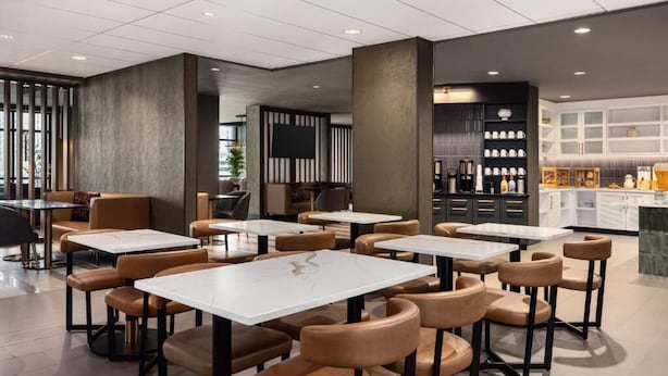 A modern restaurant interior with white marble tables and brown leather chairs, a bar area, and a kitchen visible in the background. - Hyatt place uptown (Charlotte)