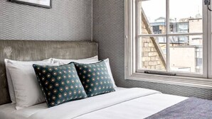 Une chambre avec un grand lit, une fenêtre donnant sur des immeubles et un mur décoré d’un motif.