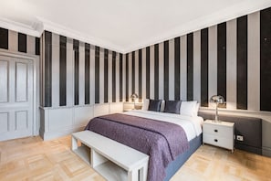 A bedroom with a bed, bedside tables, a bench, a door, and a striped wall. - Exclusive Cadogan Square Private Elevator (London)
