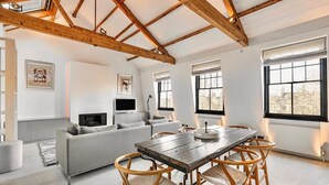 A spacious living room with a high ceiling, wooden beams, a grey sofa, a wooden dining table with chairs, and large windows. - Cadogan Square 3BR Prime Location (London)
