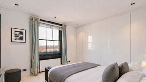 A modern bedroom with a large bed, a window with curtains, a bedside table, and a chair. - Cadogan Square 3BR Prime Location (London)