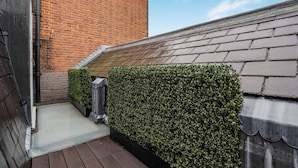 A narrow outdoor corridor with a brick wall, a wooden deck, and a hedge. - Cadogan Square Penthouse Huge Terrace AC (London)