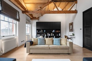 A living room with a high wooden ceiling, a beige sofa with patterned cushions, a black entertainment unit, and a white radiator. - Cadogan Square Penthouse Huge Terrace AC (London)